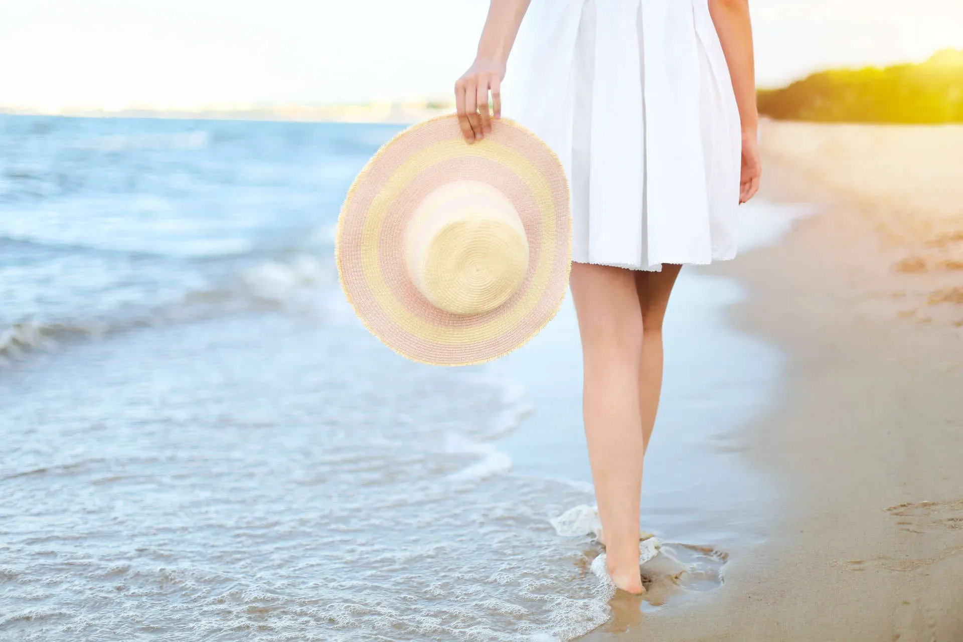 Woman walking on beach holding sun hat.