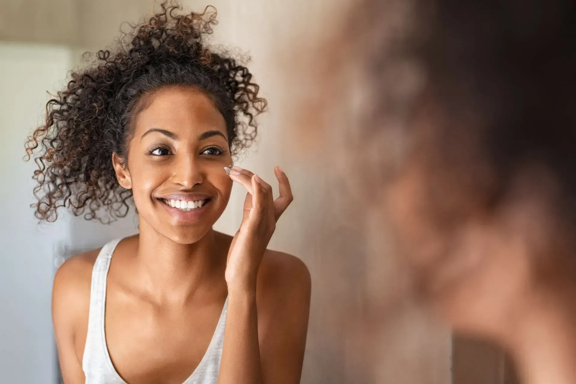 Smiling woman applying skincare in mirror reflection.